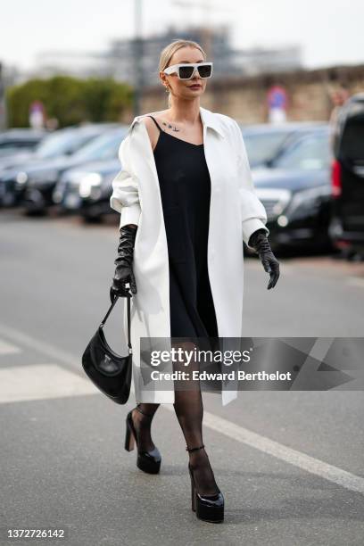 Justyna Czerniak wears white sunglasses, gold earrings, a white oversized long coat, a black V-neck / tank-top knees dress, a black shiny varnished...