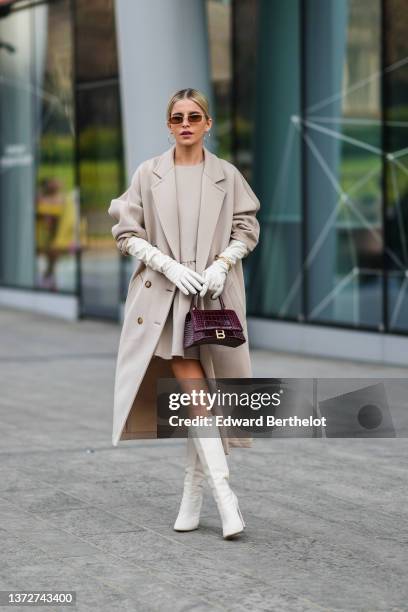 Caroline Daur wears brown sunglasses, a beige ruffled belt short dress, a beige long coat, white leather high gloves, gold large chain bracelets, a...