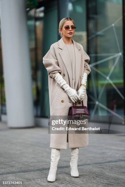 Caroline Daur wears brown sunglasses, a beige ruffled belt short dress, a beige long coat, white leather high gloves, gold large chain bracelets, a...