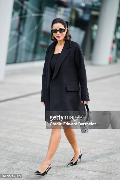 Bettina Looney wears black sunglasses, gold earrings, a black knot in the hair, a black shoulder-off short dress, a black small shiny leather belt, a...