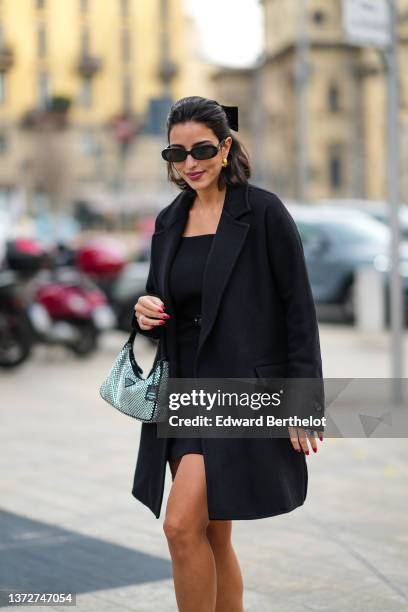 Bettina Looney wears black sunglasses, gold earrings, a black knot in the hair, a black shoulder-off short dress, a black small shiny leather belt, a...