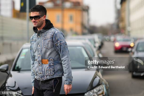 Marc Forne wears black sunglasses, silver and gold earrings, a black hoodie sweater, a blue faded denim jacket from Dolce and Gabbana, black large...