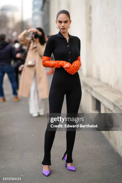 Guest wears silver earrings, a black ribbed wool belted long jumpsuit, neon orange shiny leather high gloves from Prada, purple satin pointed pumps...