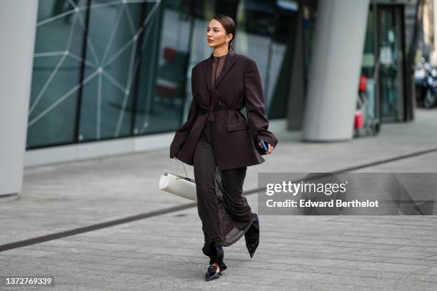 Guest wears silver earrings, a dark brown transparent long dress, a dark brown belted oversized blazer jacket, matching dark brown flared suit pants,...