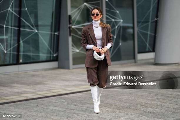 Guest wears sunglasses, gold earrings, a white ribbed wool turtleneck pullover, a dark brown and small white striped print pattern oversized blazer...