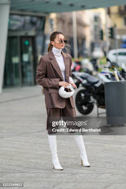 Guest wears sunglasses, gold earrings, a white ribbed wool turtleneck pullover, a dark brown and small white striped print pattern oversized blazer...