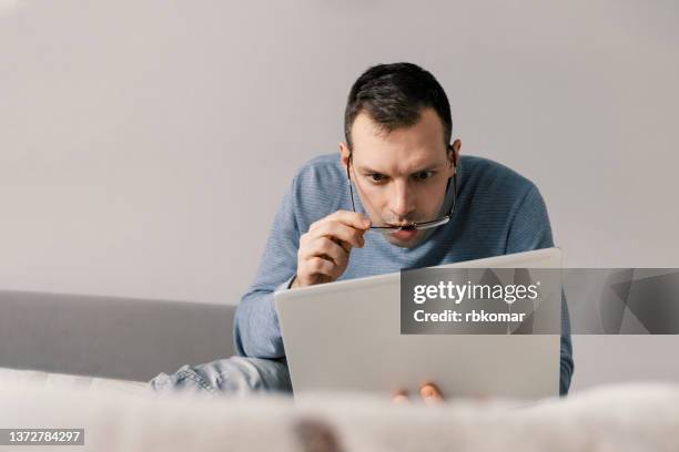 shocked astonished man in eyeglasses looking at laptop screen while sitting on sofa at home. facial expression - wow - man shock stockfoto's en -beelden