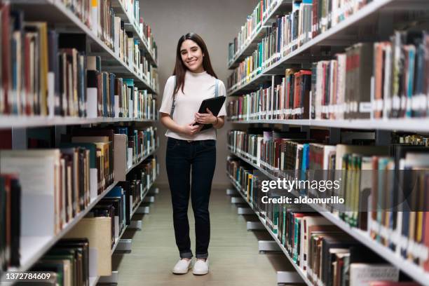 friendly female student standing between bookshelves in library - choice student stockfoto's en -beelden