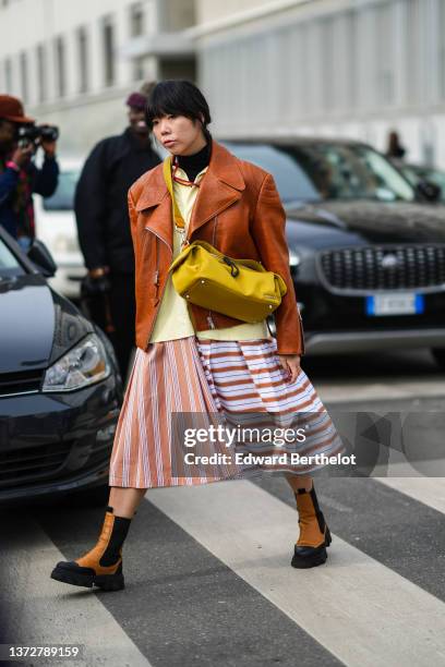 Guest wears silver earrings, a black turtleneck pullover, red glasses, a yellow shirt, a camel shiny leather zipper oversized jacket, a brown and...