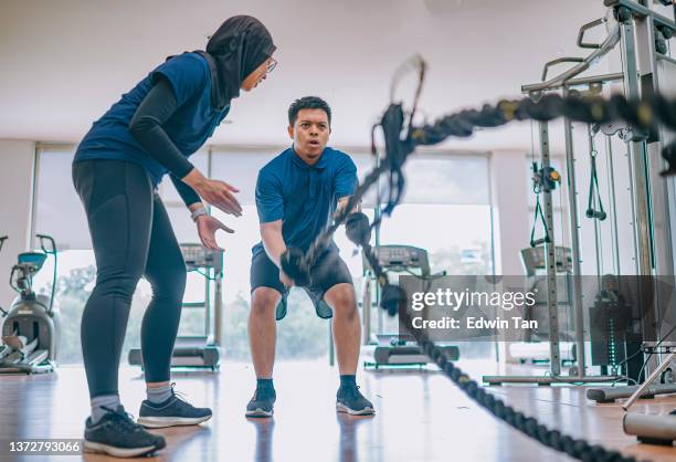 asiatisch-malaiische personal trainerin ermutigt asiatischen malaiischen mann, der herausforderndes kampfseil im fitnessstudio übt - hiit stock-fotos und bilder