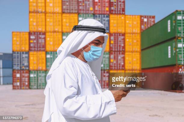 arab businessman with face mask using mobile phone standing outside on a large commercial dock during pandemic - coronavirus uae bildbanksfoton och bilder
