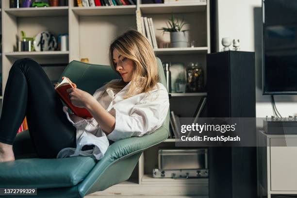 a beautiful blonde woman sitting in her armchair and reading a book at home - casual woman pensive side view stockfoto's en -beelden