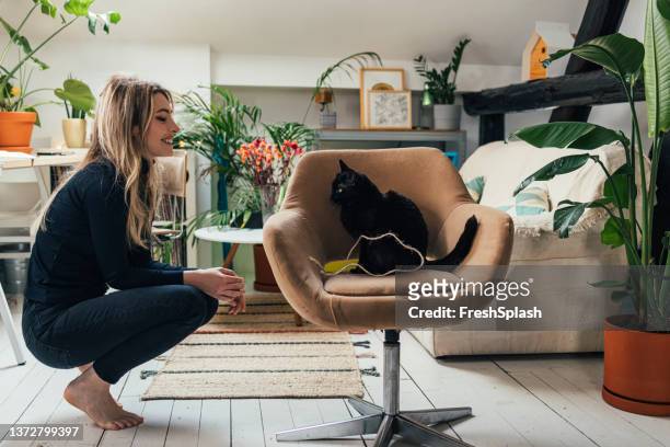 una vista laterale di una bella donna bionda che sorride mentre guarda il suo simpatico gatto nero seduto sulla sua sedia da ufficio a casa - black and white cat foto e immagini stock