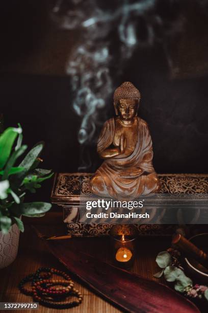 low key still life of a golden buddha figurine with a smoking incense stick, a lighted candle, a singing bowl, prayer beads and a green plant in a pod - low key imagens e fotografias de stock