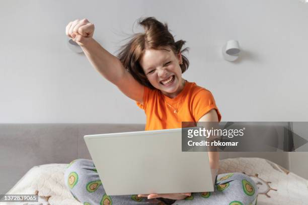 excited happy teen girl student winner celebrating victory raising hand while using laptop sitting in lotus pose on bedroom. received great news online looking at computer. success concept, wireless technology and fast internet speed - one teenage girl only ストックフォトと画像