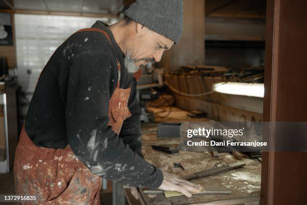 side view of artisan carpenter working in his workshop wearing very dirty and old red overalls and a gray cap on a customer's order. - dirty construction worker stock pictures, royalty-free photos & images