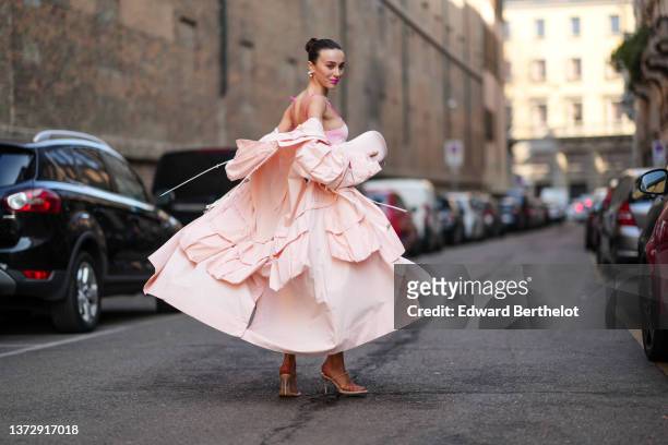 Mary Leest wears silver large earrings, a pale pink tank-top tube knees with embroidered pale pink ruffled / pleated midi dress, a pale pink...