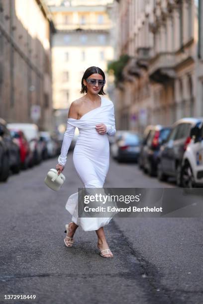 Alexandra Pereira wears sunglasses, a white asymmetric shoulder / long sleeves / wrap chest / ruffled ankle long dress, a white latte zipper handbag,...