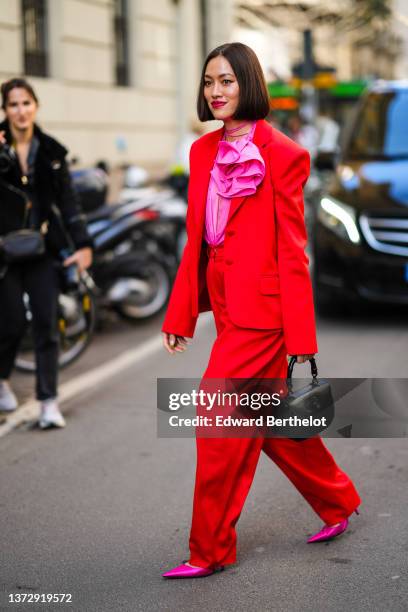 Tiffany Hsu wears a neon pink ruffled blouse, a neon red blazer jacker, matching red large suit pants, a black shiny leather handbag, neon pink satin...