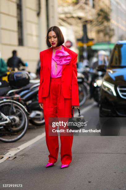 Tiffany Hsu wears a neon pink ruffled blouse, a neon red blazer jacker, matching red large suit pants, a black shiny leather handbag, neon pink satin...