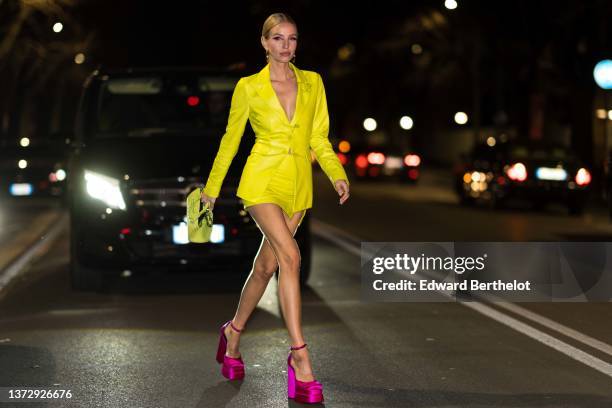 Leonie Hanne wears gold large earrings from Versace, a yellow shiny leather belted blazer jacket, a yellow shiny leather high waist slit short skirt,...