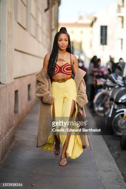 Ellie Delphine wears gold earrings, gold chain pendant necklaces, a red / yellow braided macrame halter-neck / cropped top, a brown oversized long...