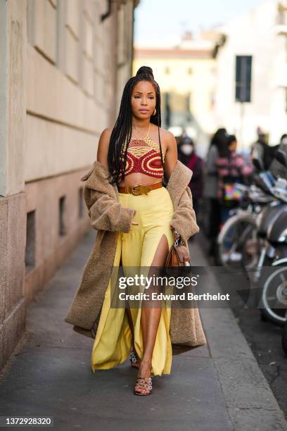 Ellie Delphine wears gold earrings, gold chain pendant necklaces, a red / yellow braided macrame halter-neck / cropped top, a brown oversized long...