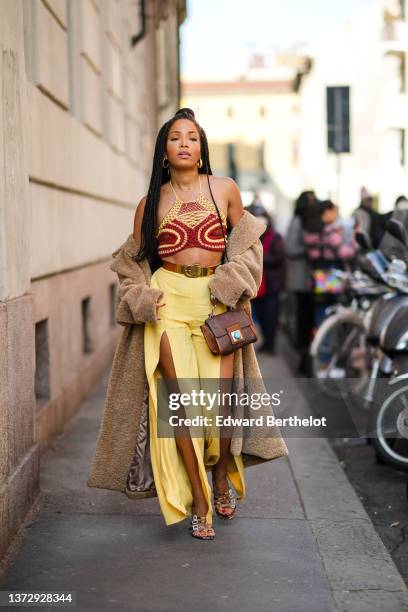 Ellie Delphine wears gold earrings, gold chain pendant necklaces, a red / yellow braided macrame halter-neck / cropped top, a brown oversized long...