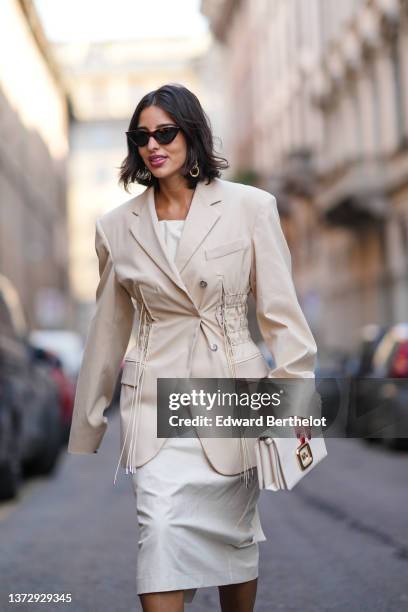 Bettina Looney wears sunglasses, gold earrings, a white shoulder-off blouse, a beige ruffled / epaulets blazer jacket, matching beige leather clutch,...