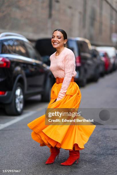 Guest wears silver earrings, a pale pink cut-out print pattern wool pullover, a camel shiny leather large belt, an orange ruffled / slit long skirt,...