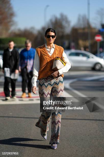 Guest wears sunglasses from Ray Ban, a white shirt, a camel braided wool sleeveless pullover, diamond earrings, a beige / yellow / camel shiny...