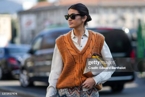 Guest wears sunglasses from Ray Ban, a white shirt, a camel braided wool sleeveless pullover, diamond earrings, a beige / yellow / camel shiny...