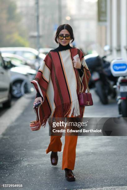 Guest wears black sunglasses, a black turtleneck pullover, a red / white / black striped print pattern fringed cloak, a burgundy shiny leather...