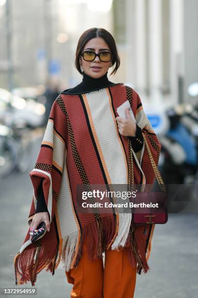 Guest wears black sunglasses, a black turtleneck pullover, a red / white / black striped print pattern fringed cloak, a burgundy shiny leather...
