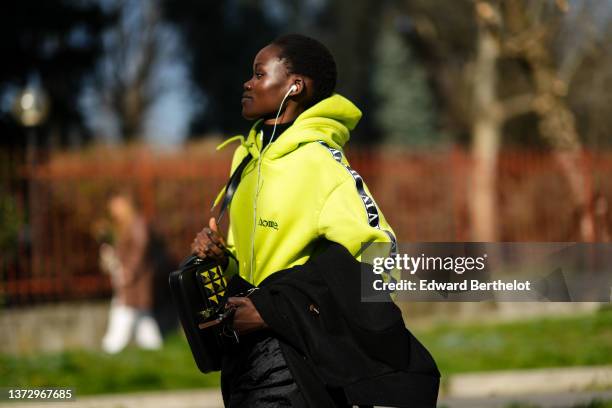 Guest wears a neon yellow and black print pattern strap hoodie sweater from Home, a black coat, a black shiny leather shoulder bag, black pants, a...