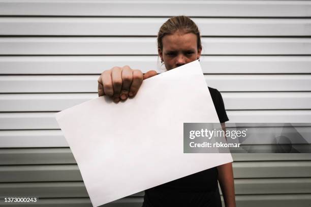 portrait of an upset angry adult girl showing blank board and looking at camera against a striped wall outdoors. copy space for text. activist with empty banner and protest concept - protestor portrait stock pictures, royalty-free photos & images