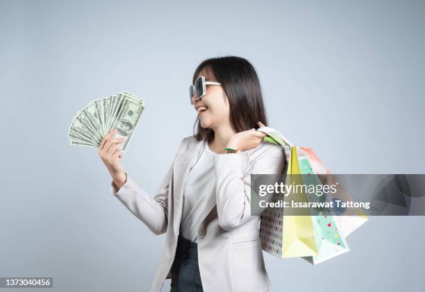 photo of cheerful elegant young woman holding fan of money and colorful shopping bags, looking aside, isolated on gray background - bônus - fotografias e filmes do acervo