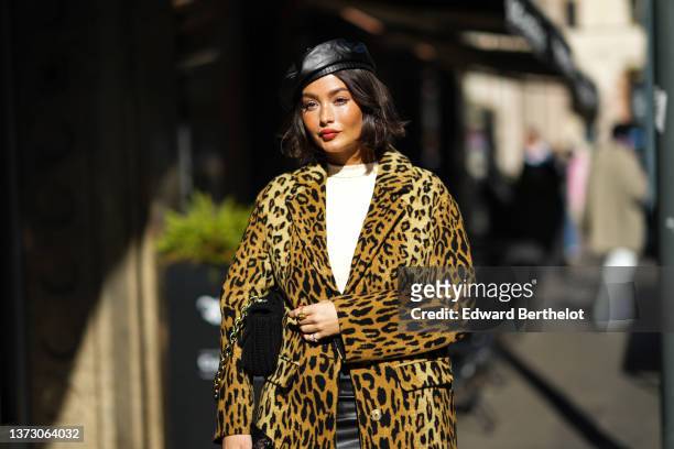 Taylor Lashae wears a black shiny leather beret, a white high neck pullover, a black shiny leather short skirt, a brown and black leopard print...