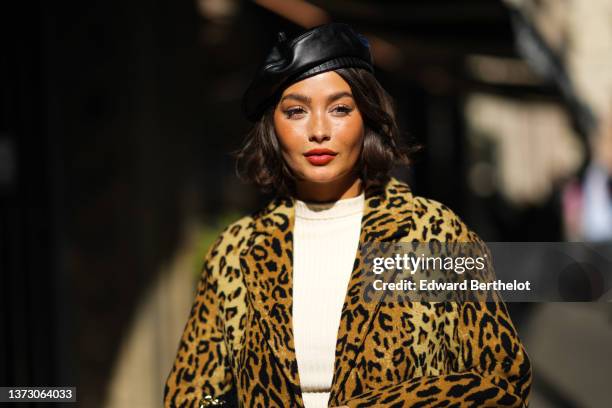 Taylor Lashae wears a black shiny leather beret, a white high neck pullover, a black shiny leather short skirt, a brown and black leopard print...