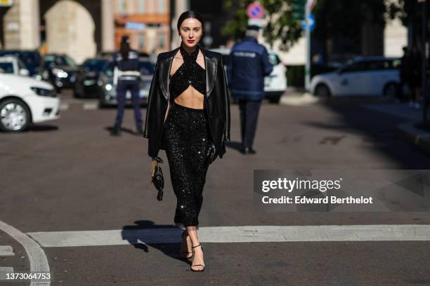 Mary Leest wears silver earrings, a black shiny leather blazer jacket, a black sequined crossed neck / cropped top, a matching black shiny...