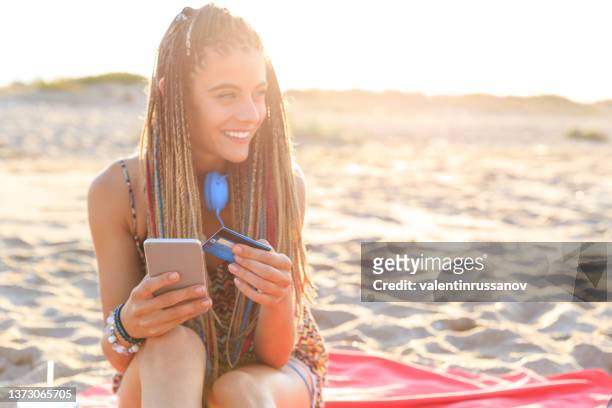 beautiful smiling girl with braided hair sitting on the beach with her mobile phone  at sunset, holding a credit card and making online payments, online shopping and digital purchases, during her vacation - beach towel stockfoto's en -beelden