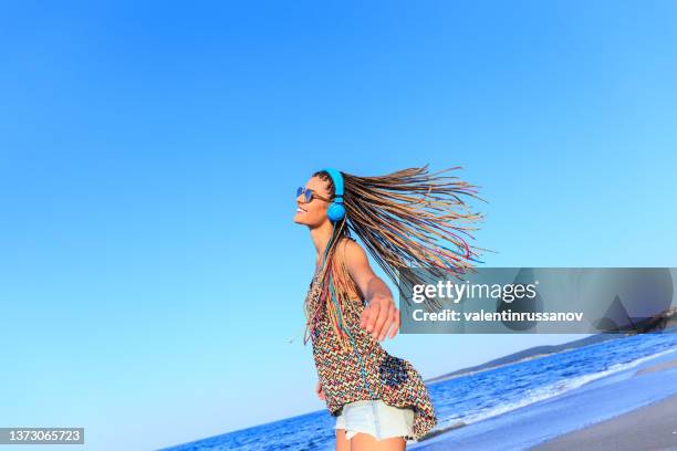 smiling and joyful young woman with braided hair and blue headphones listening to music and dancing on the beach. - beach music stock pictures, royalty-free photos & images