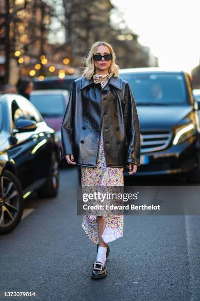 Candela Novembre wears black sunglasses, a white with yellow / purple / pink flower print pattern high neck / asymmetric midi dress, a black shiny...