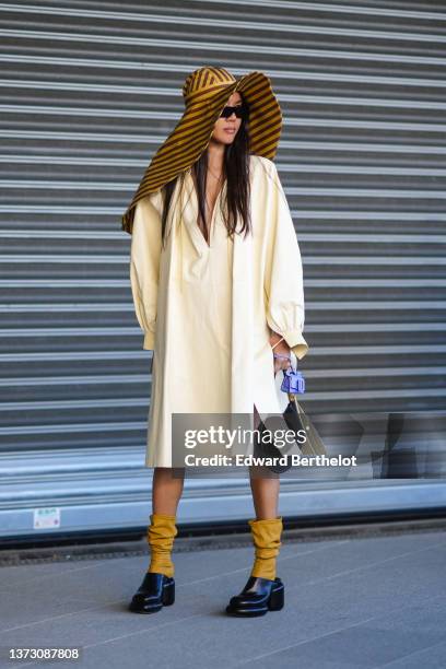 Guest wears a yellow mustard and dark brown striped print pattern oversized hat, black sunglasses, a pale yellow oversized long shirt dress, a black...
