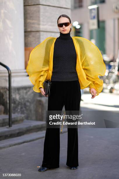 Guest wears black and gold sunglasses, a black braided wool turtleneck pullover, yellow ribbed tulle oversized ruffled bat sleeves, black velvet...