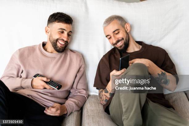 two friends watching something on the mobile phone while relaxing lying on a sofa at home. - bromance stock-fotos und bilder