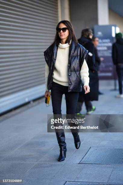 Guest wears black sunglasses, a white latte ribbed oversized high neck pullover, a black shiny quilted sleeveless oversized coat, black skinny denim...