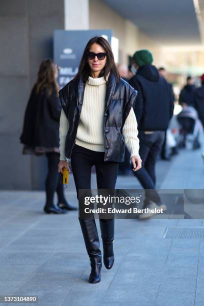 Guest wears black sunglasses, a white latte ribbed oversized high neck pullover, a black shiny quilted sleeveless oversized coat, black skinny denim...