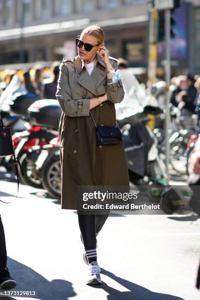 Guest wears black sunglasses, a white turtleneck pullover, a khaki and gray bicolored long trench coat, a black shiny leather WOC Classique crossbody...