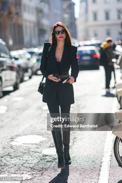 Guest wears black sunglasses, a black shiny leather accordion shoulder bag, a black V-neck t-shirt, a black long blazer jacket, a black shiny leather...